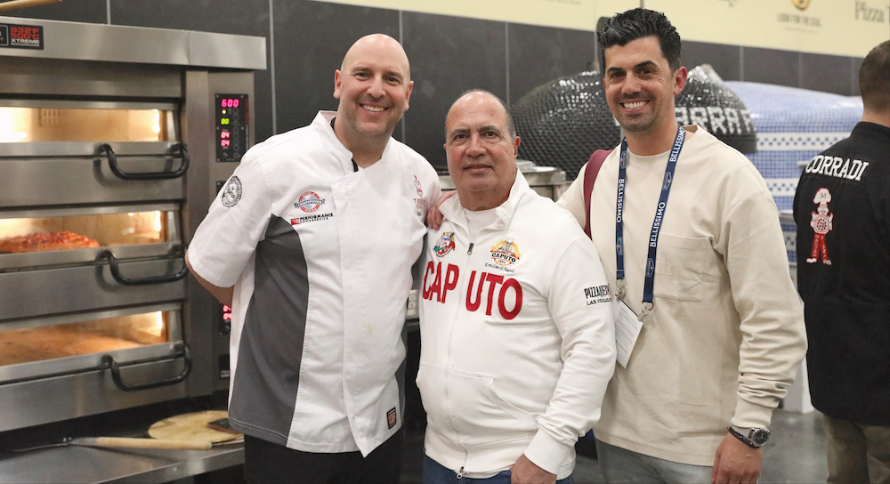 This photo shows Chris Decker in a white chef coat, John Arena in a white Caputo zip-up sweater and Michael Vakneen standing next to a deck oven.