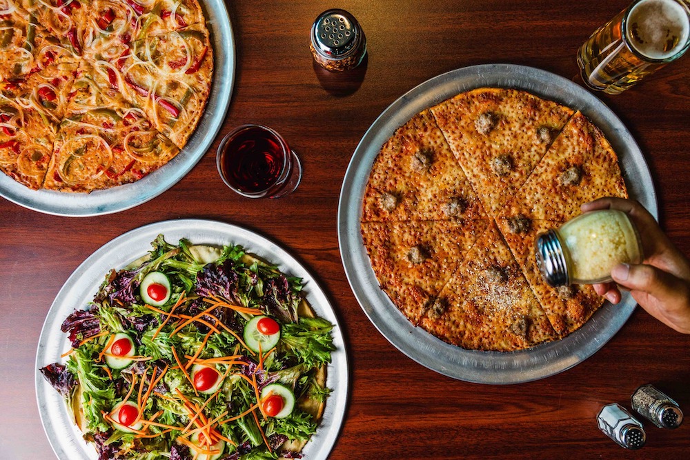 this photo shows three pizzas from Colony Grill with a Parmesan cheese container hovering over one of them.