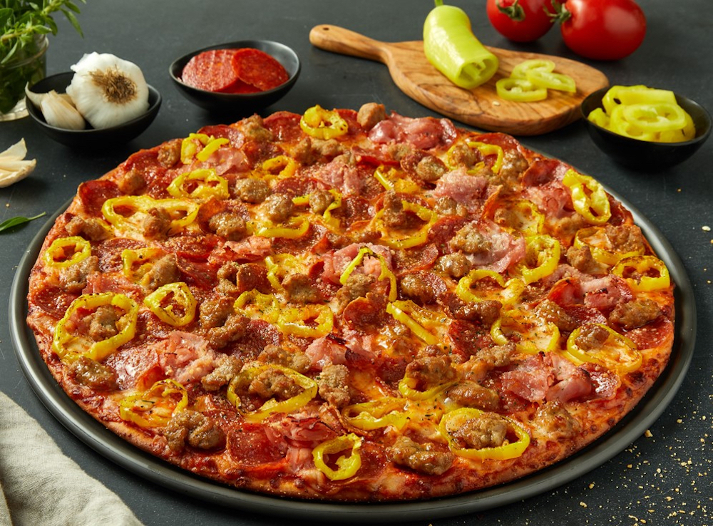 This photo shows a tray with a whole pizza topped with pepperoni, sausage and yellow peppers, with small bowls of ingredients - pepperoni, onion and jalapeños - in the background.