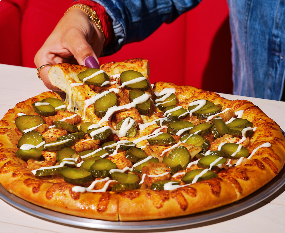 this photo shows a woman's hand removing a slice of a pizza topped with pickle slices, breaded chicken breast and white onions with a drizzle of buttermilk ranch sauce