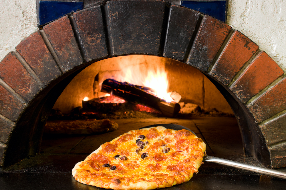 This photo shows a pepperoni and olive pizza coming out of a wood burning oven.