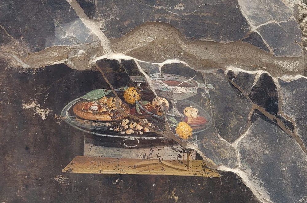 this is a photo of an ancient still-life fresco found in Pompeii. It depicts what appears to be a pizza-like flatbread and various fruits, such as pomegranates.