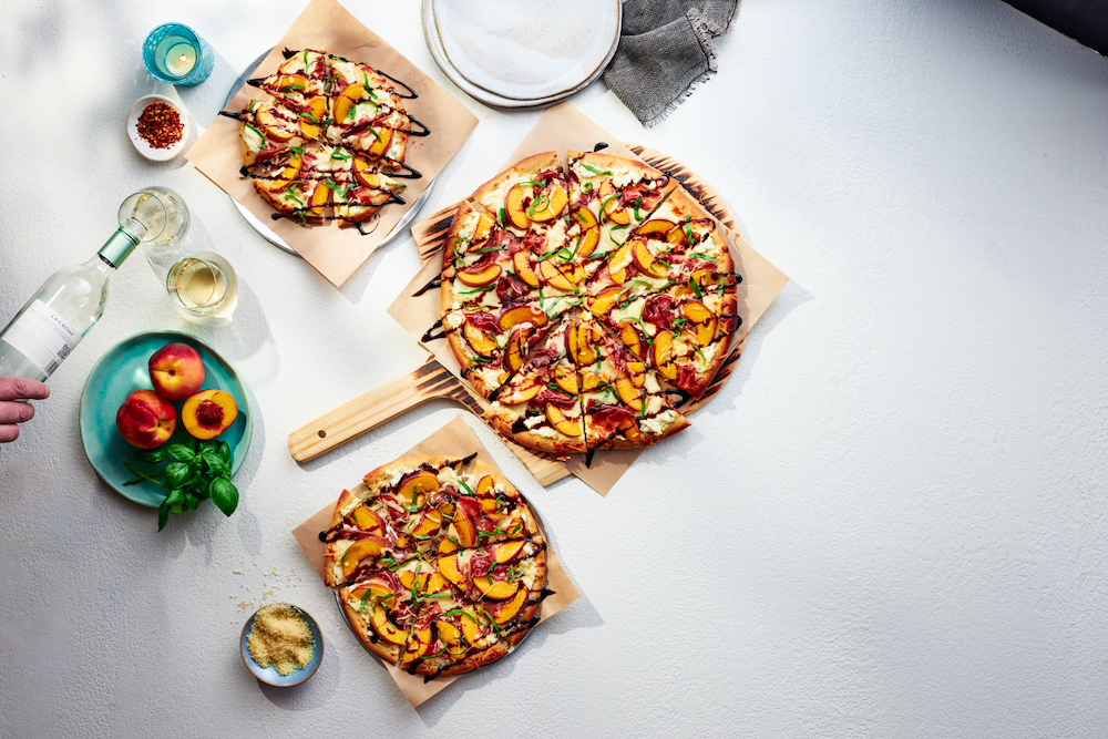 this photo shows three peach-topped pizzas against a white background while a bottle of wine is being poured into two glasses