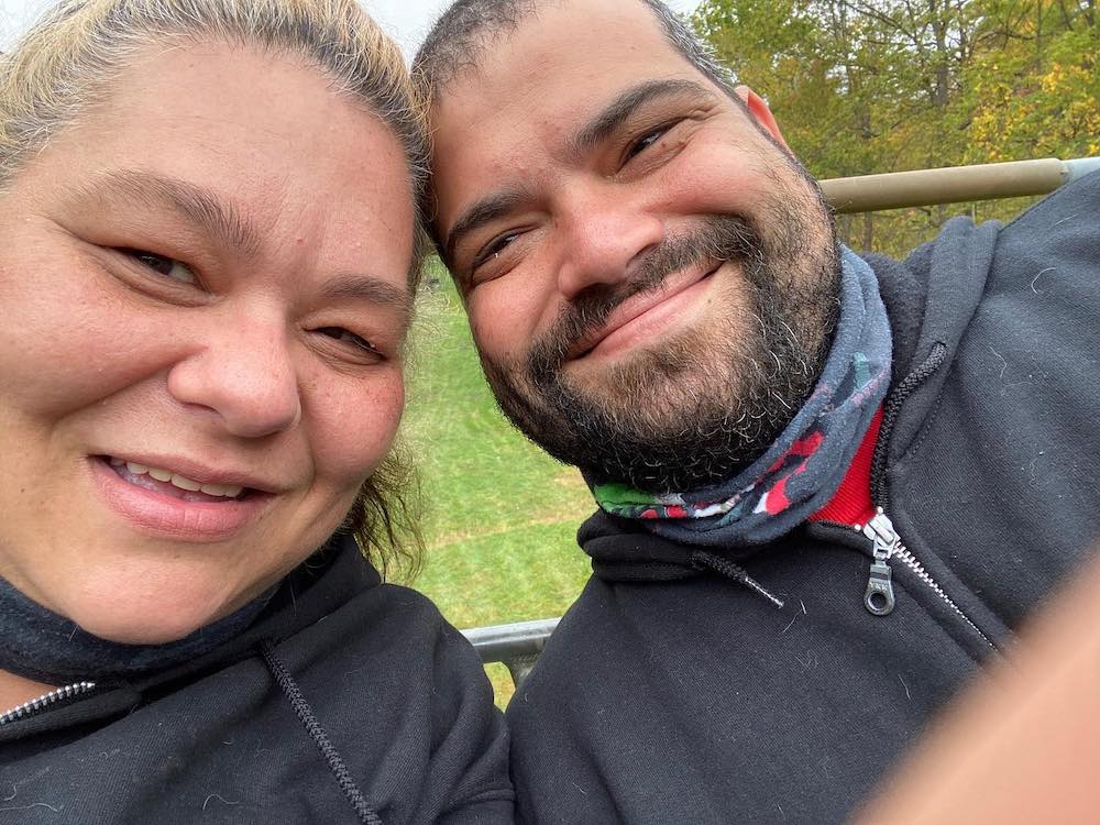 This selfie-style photo shows a close-up of Carly and Tom Cinelli, smiling in an outdoor setting with their heads together