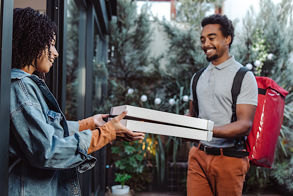 this photo shows a young African-American delivery man holding two pizza boxes out for a young African-American woman customer at her doorstep.