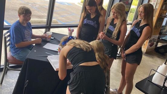 This photo shows Wes Besancon seated at a table while a group of high school cheerleaders gather around to sign his yearbook.