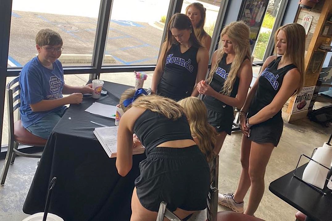 This photo shows Wes Besancon seated at a table while a group of high school cheerleaders gather around to sign his yearbook.