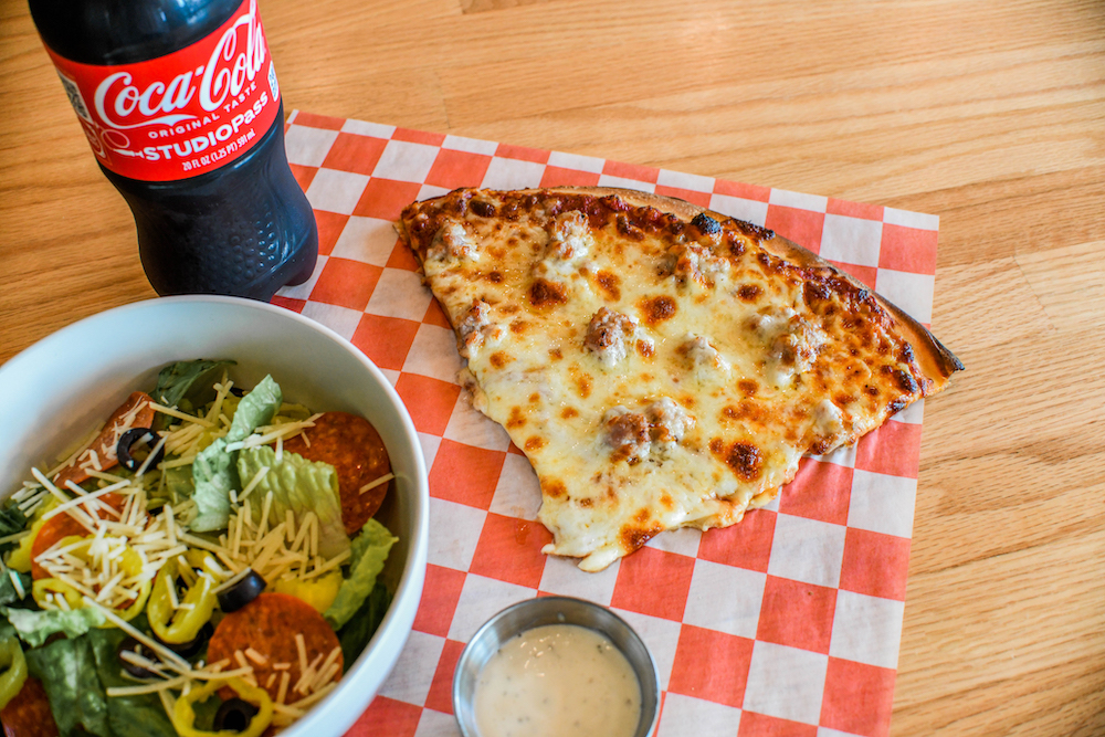 This photo shows a large pizza slice with a salad, a mini-cup of dressing and a bottle of Coke