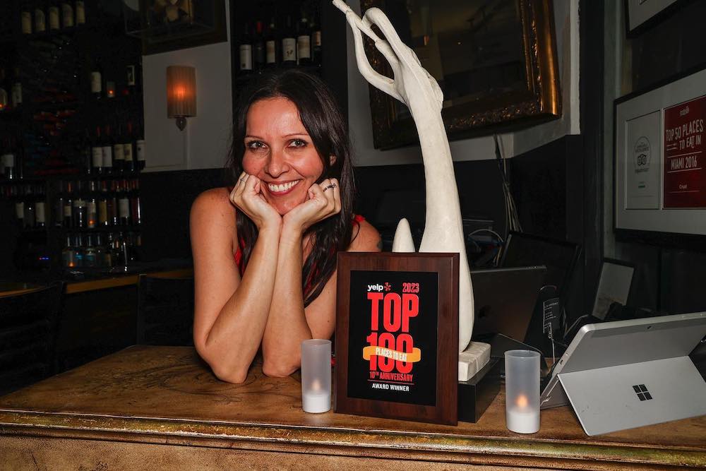 This photo shows a warmly smiling Anita Kovaceski, her face resting in her hands, with a plaque noting that Crust made Yelp's list of the Top 100 Places to Eat for 2023.