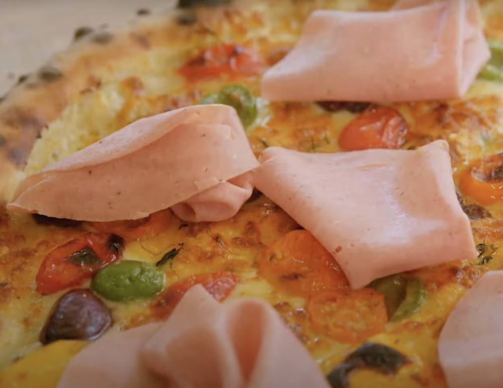 this photo shows a close-up of a pizza topped with curls of bologna, roasted tomatoes, mozzarella, olives and a creamy garlic sauce.