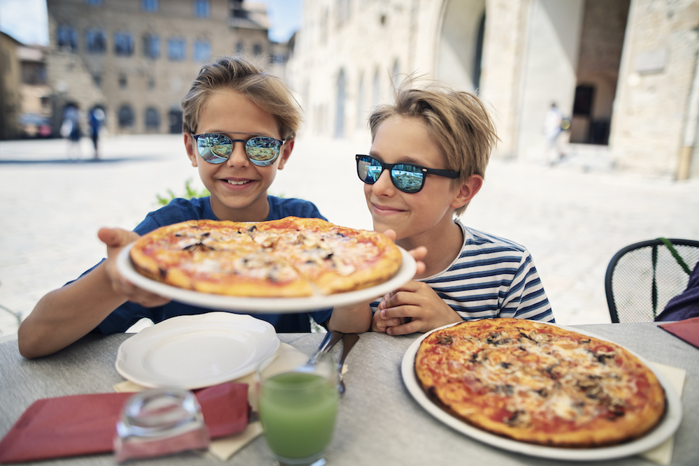 This photo shows two cute boys in sunglasses dining outdoors in the the beautiful Italian town of Volterra. One of the boys is holding up a pizza while the other boy peers in for a closer look. There's a second pizza on the table in front of the second boy.