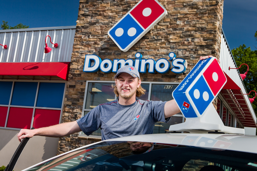 This photo shows a smiling young man, with longish sandy-colored hair and a mustache and beard, standing next to a Domino's delivery car with the Domino's sign on top.