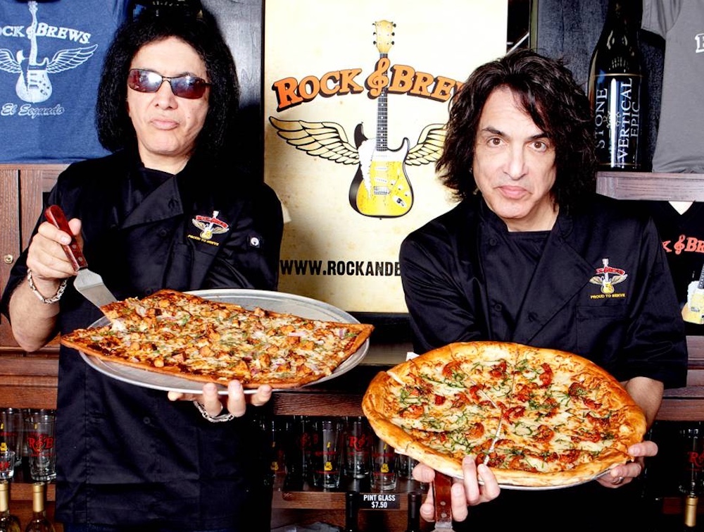 This photo shows Gene Simmons and Paul Stanley standing in front of a Rock & Brews banner, each holding a large pizza on a tray.