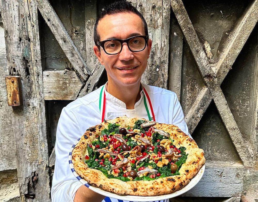 This photo shows Gino Sorbillo, in a white chef coat and wearing glasses, holding up the Pompeii Pizza, topped with spinach, anchovies, pomegranate, olives, walnuts and other ingredients.