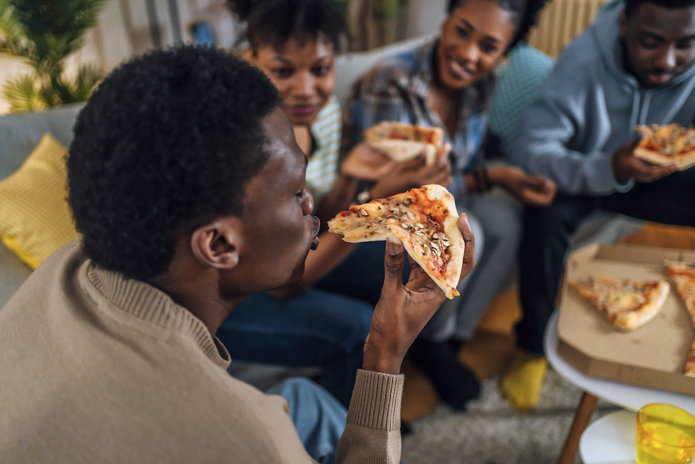 This photo shows a group of young, cheerful, African-American friends sitting in a living room and eating pizza, with the man in the foreground having happily just taken a bite out of a slice.
