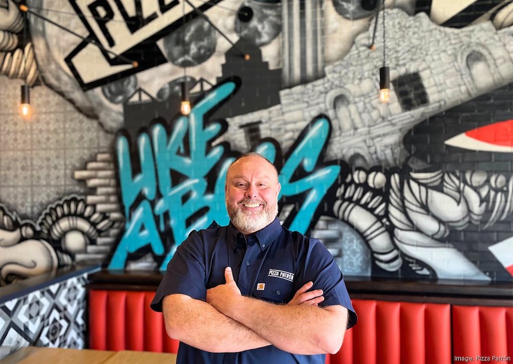 This photo shows Guy Carney, with thinning hair and a grey and dark beard, standing with his arms crossed and smiling in front of a mural at a Pizza Patrón location.