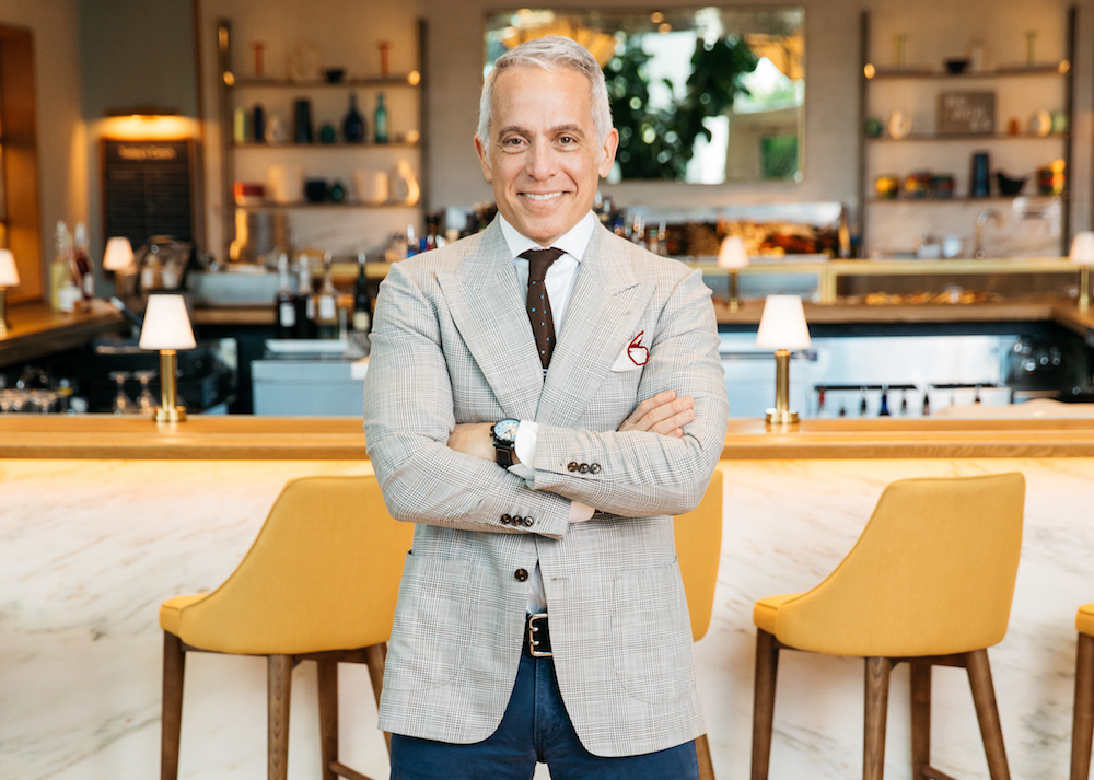 This photo shows Geoffrey Zakarian, with gray hair and wearing a light-colored sports coat with a tie, standing in a restaurant with his arms crossed and smiling for the camera.