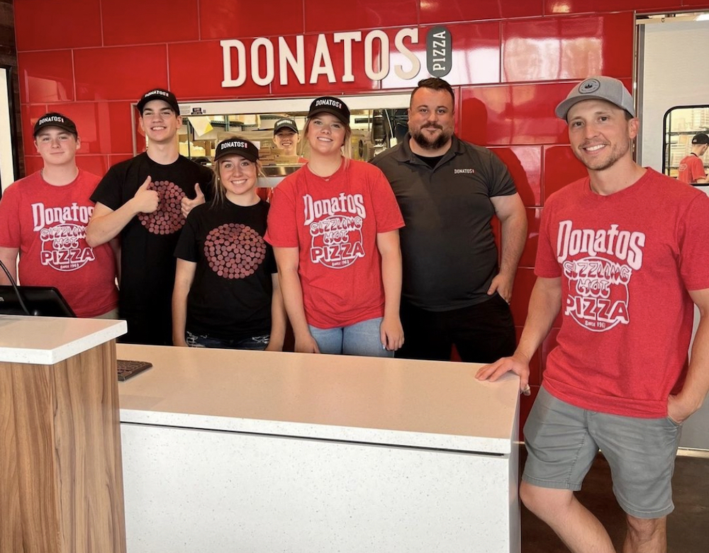 this photo shows a group of employees in red branded Donatos t-shirts standing in a newly opened location and smiling for the camera.