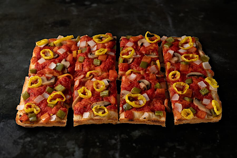 This photo shows a gluten-free Detroit-style pizza topped with pepperoncini, mushrooms, green bell peppers and onions, set against a charcoal-gray backdrop.