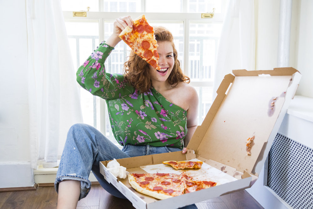 this photo shows Julia Chebotar, with red hair and wearing an off-the-shoulder green blouse, holding up a slice of pepperoni pizza from a delivery box.