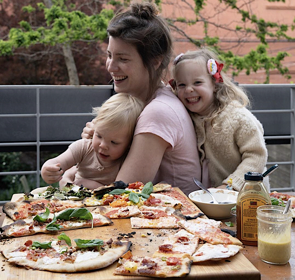 This photo shows Lauren Grimm at a table festooned with various pizzas, while two little girls hang onto her. Everyone is smiling and laughing.