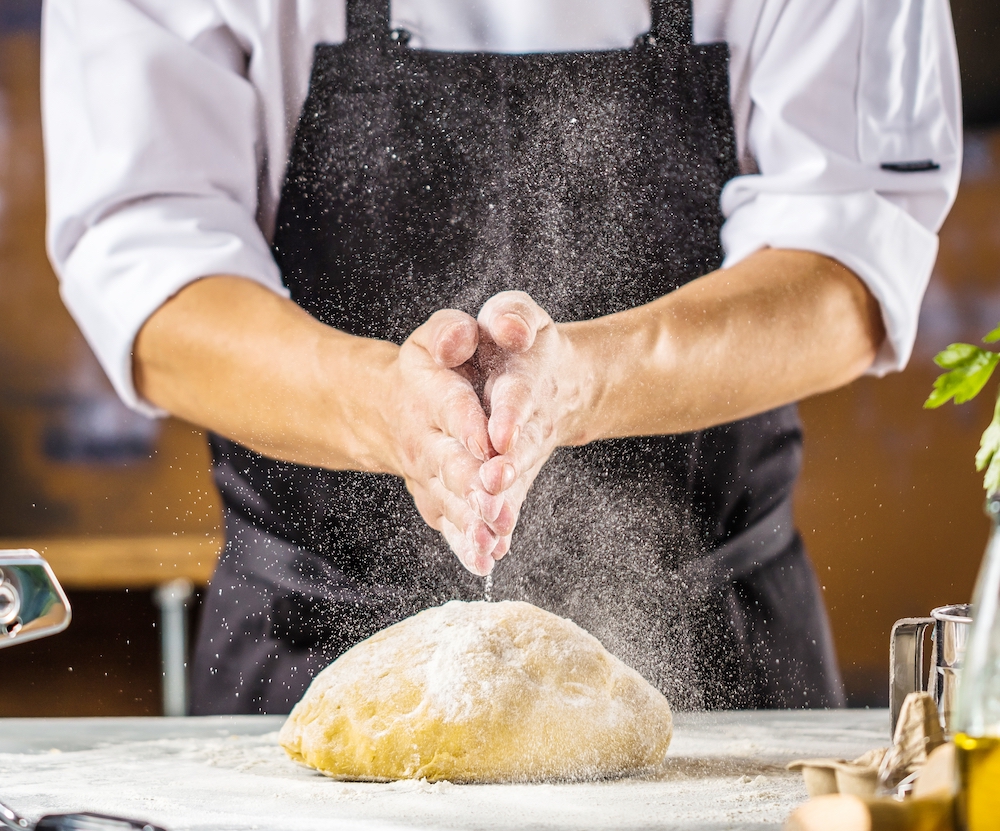 This shows a male chef, seen only from the chest down, prepping a ball of pizza dough on a table, with his hands pressed together over the dough.