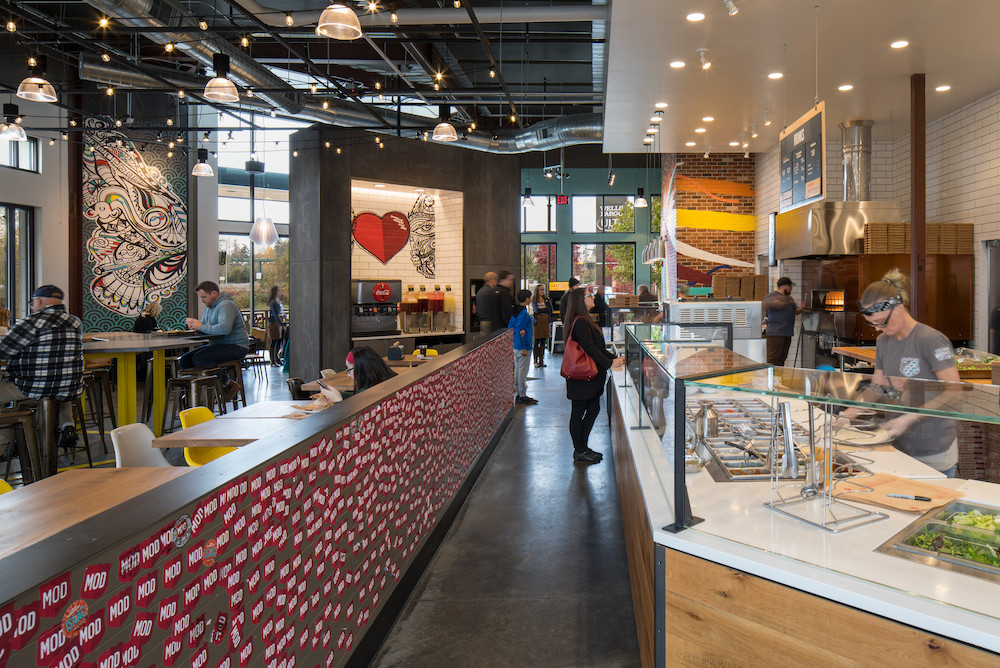 This photo shows the interior of a MOD Pizza location, with employees topping pizzas in the line, a customer looking at the menu board and other customers eating at tables.