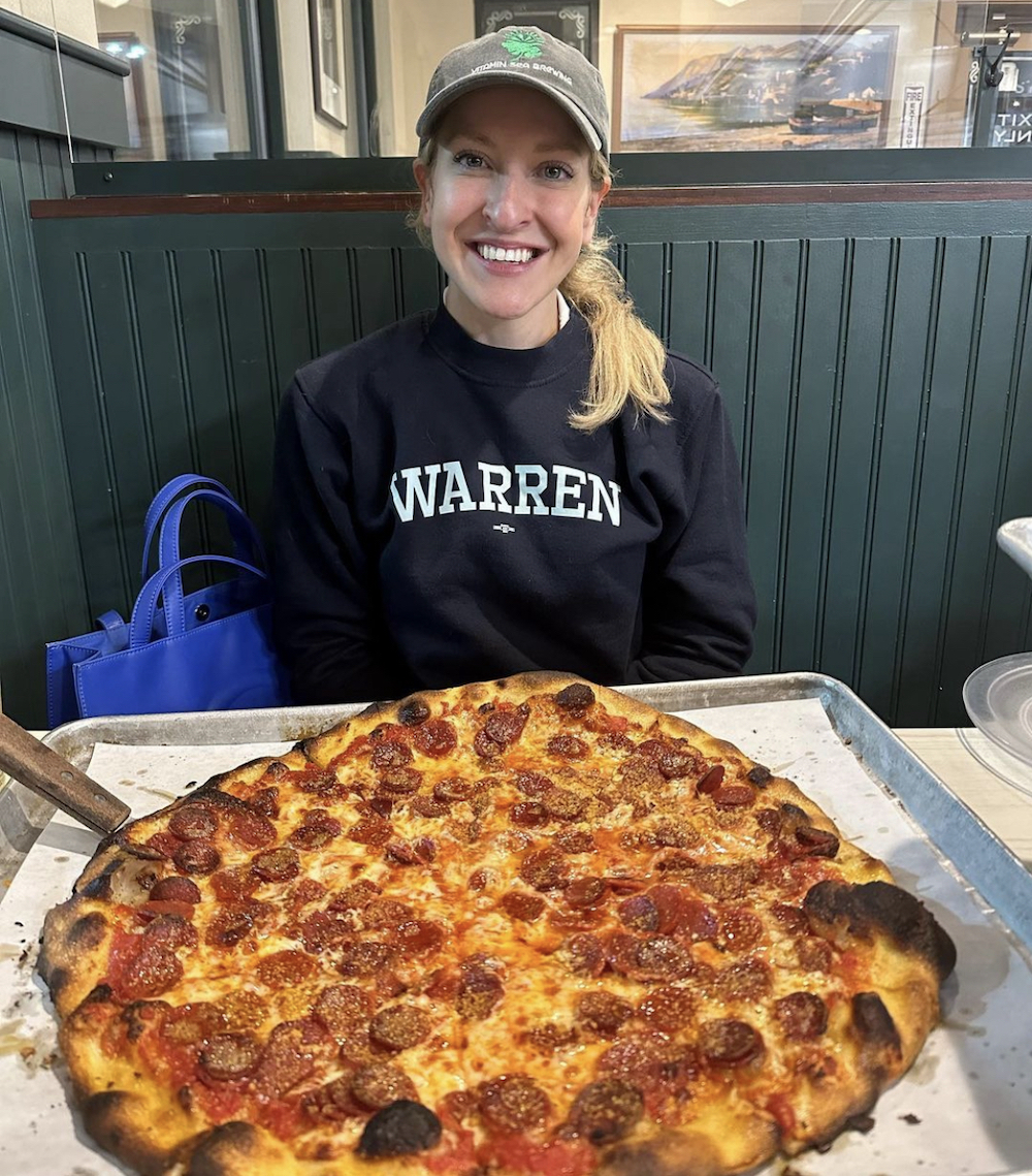 This photo shows a smiling Monica Wojnilo in a pizzeria, wearing a cap with a long blonde ponytail, sitting at a table in front of a large pepperoni pizza