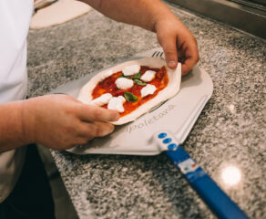 This photo shows a man's hands placing an uncooked Margherita pizza on an aluminum pizza peel.