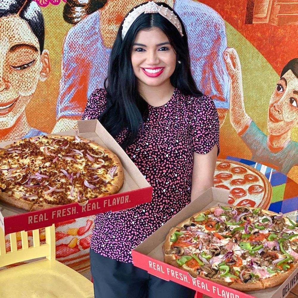 This photo shows a smiling Latino woman holding two boxed pizzas and standing before a mural at a Pizza Patrón location.