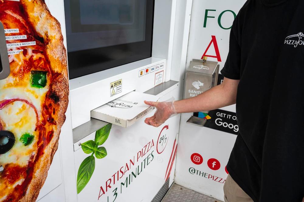 This photo shows a man removing a boxed pizza from the dispensing slot of a PizzaForno pizza vending machine.