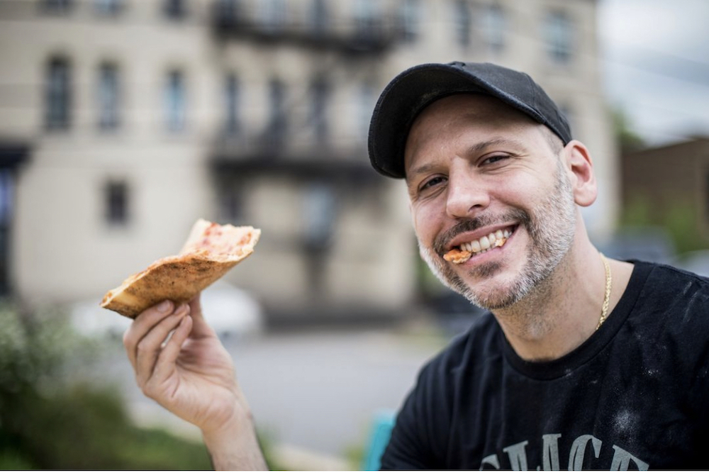 This photo, shot outdoors, shows Rico Lunardi holding up a slice and smiling, with a piece of pizza sticking out between his teeth.