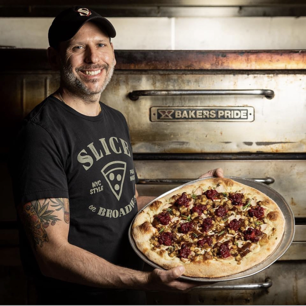 This photo shows a smiling Rico Lunardi holding a pizza in front of his oven. He's wearing a black cap and a Slice on Broadway t-shirt.