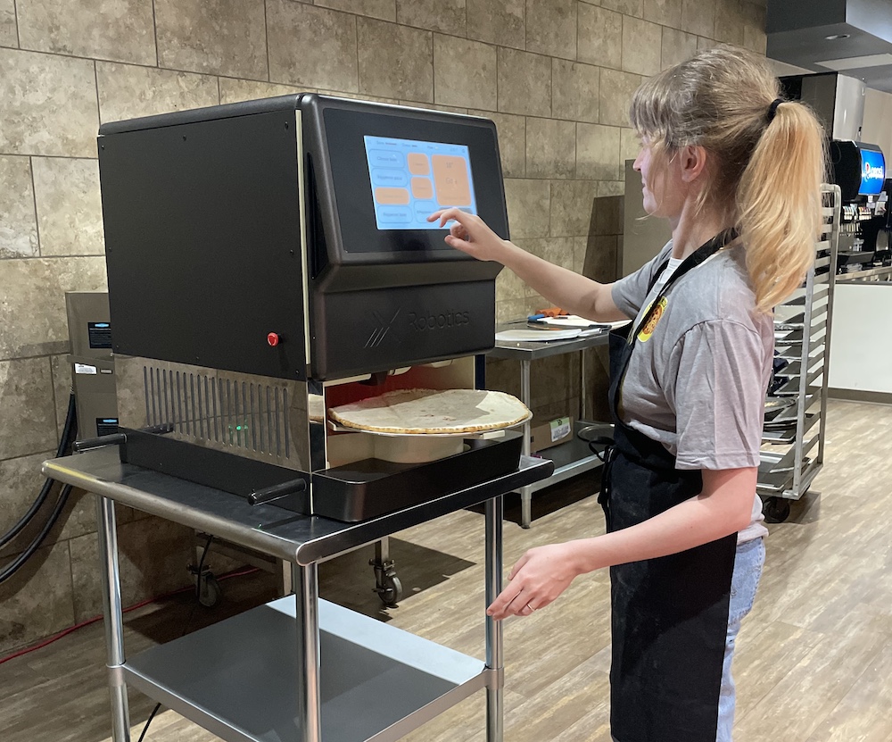 This photo shows a young woman with a blonde ponytail pressing buttons on the xRobotics xPizza Cube, a pizza-making robot.