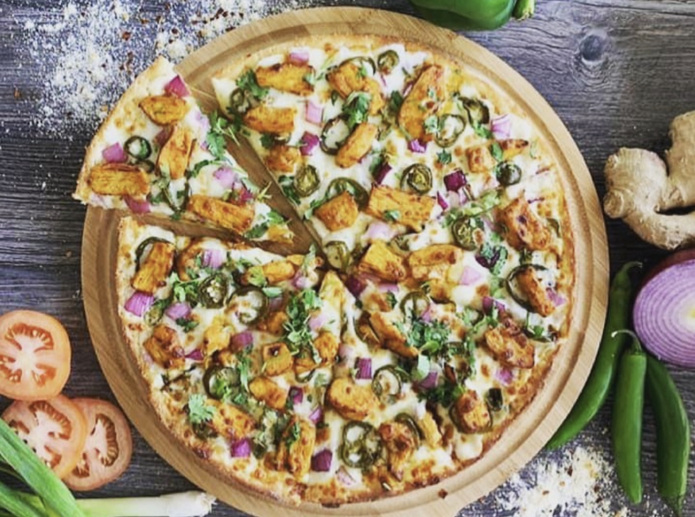 This photo shows the Achari Chicken Pizza, featuring Achari Chicken Breast, diced mozzarella cheese, red onions, spicy jalapenos, garlic, ginger, and green chilies, garnished with fresh cilantro and green onions