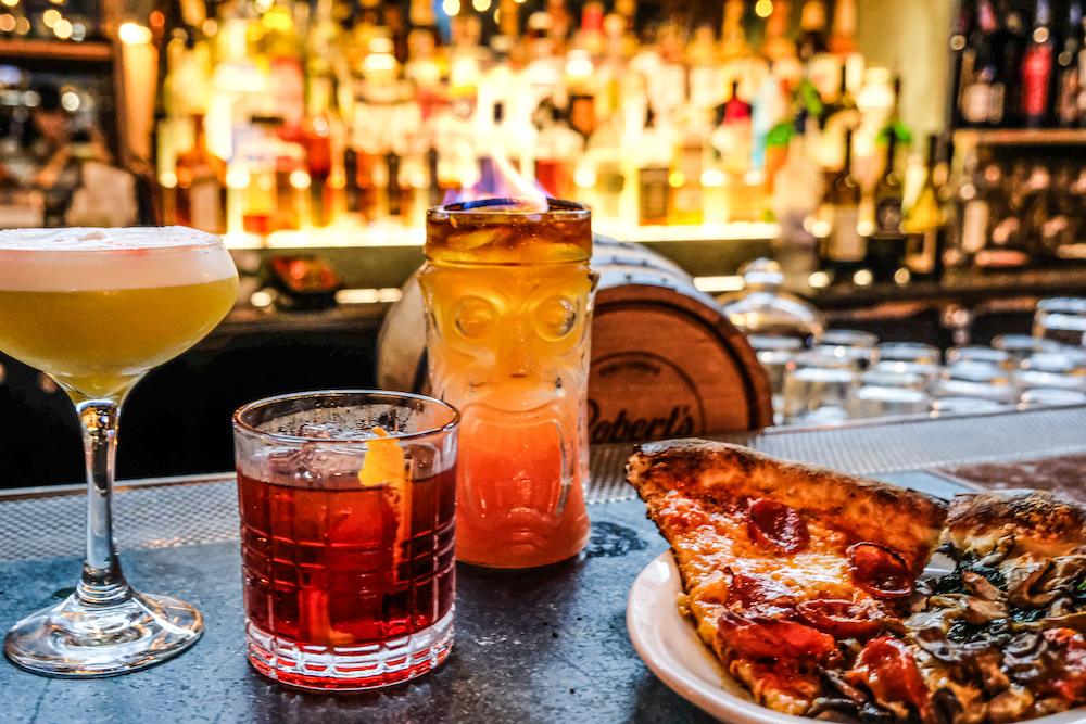 This warmly lit photo shows three cocktails alongside a slice of pepperoni pizza