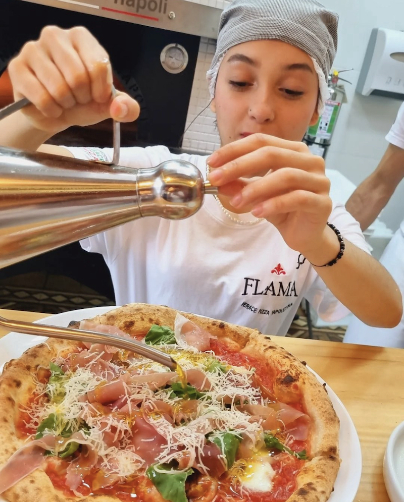 this photo shows a young woman getting ready to drizzle what appears to be olive oil on a pizza topped with prosciutto, some type of leafy vegetable and shredded fresh cheese, perhaps Parmesan.