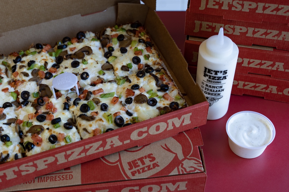 This photo shows a Detroit-style pizza in a branded Jet's box next to a bottle of Ranch dressing.