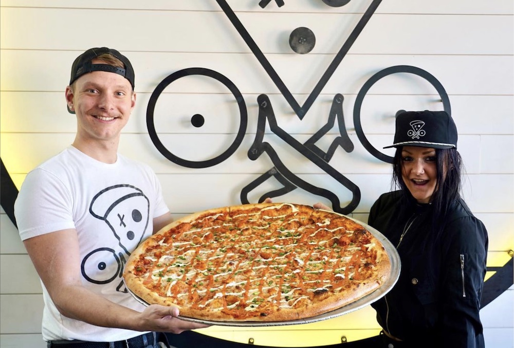 This photo shows a young man with a mustache and backwards cap posing with a giant pizza along with a young woman wearing a PIE.ZAA logoed cap, both standing in front of the PIE.ZAA logo.