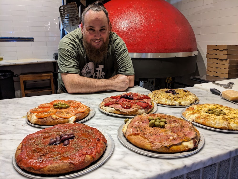 Photo shows pop-up pizza master Fernando Greco standing over six distinctly Argentine pizzas.