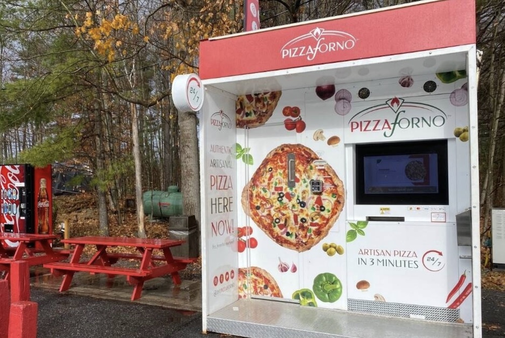 This photo shows a colorfully branded PizzaForno machine next to a red park bench in what looks to be a park with trees in the background.