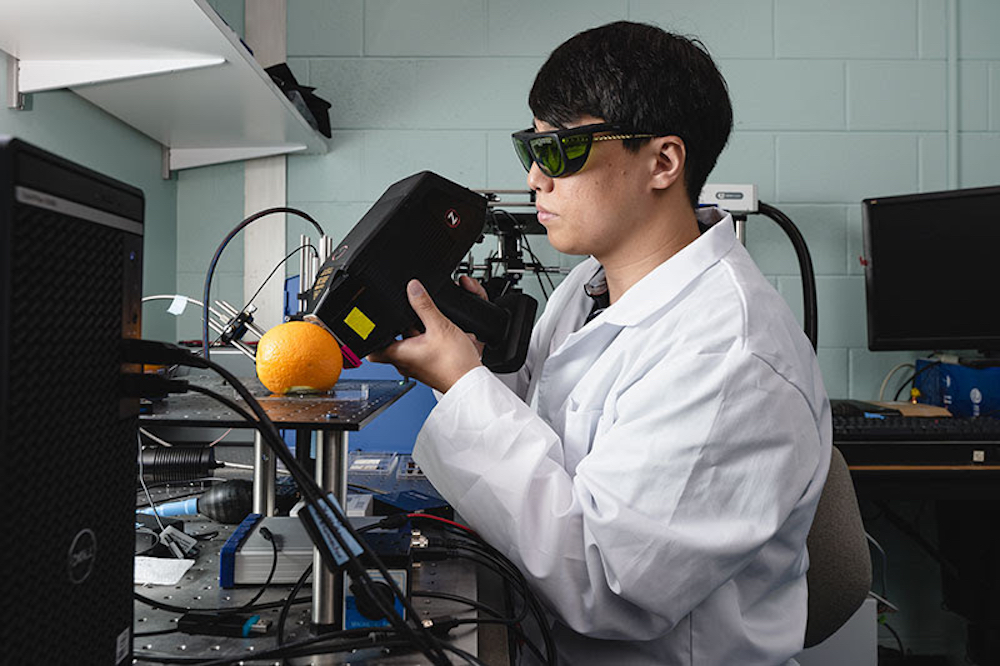 This photo shows a young man with dark hair and dark laboratory glasses peering at an orange through a portable spectroscopy instrument.