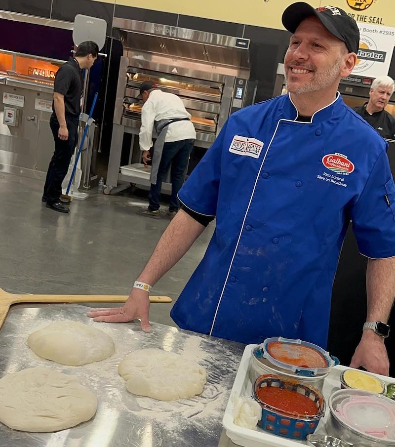 This photo shows Rico Lunardi, wearing a cap and a blue chef coat, at a pizza-making competition with a selection of dough balls and ingredients on a table in front of him.