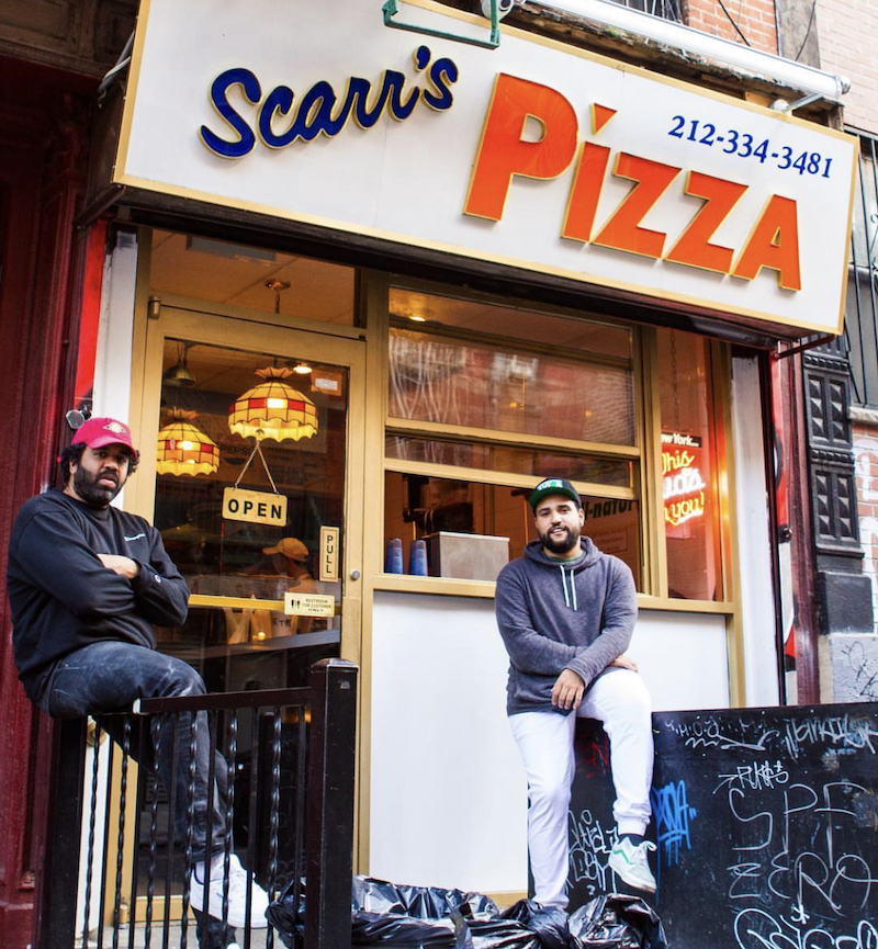 This photo shows two Black men, both sporting beards and wearing caps, sitting in front of the exterior sign for Scarr's Pizza