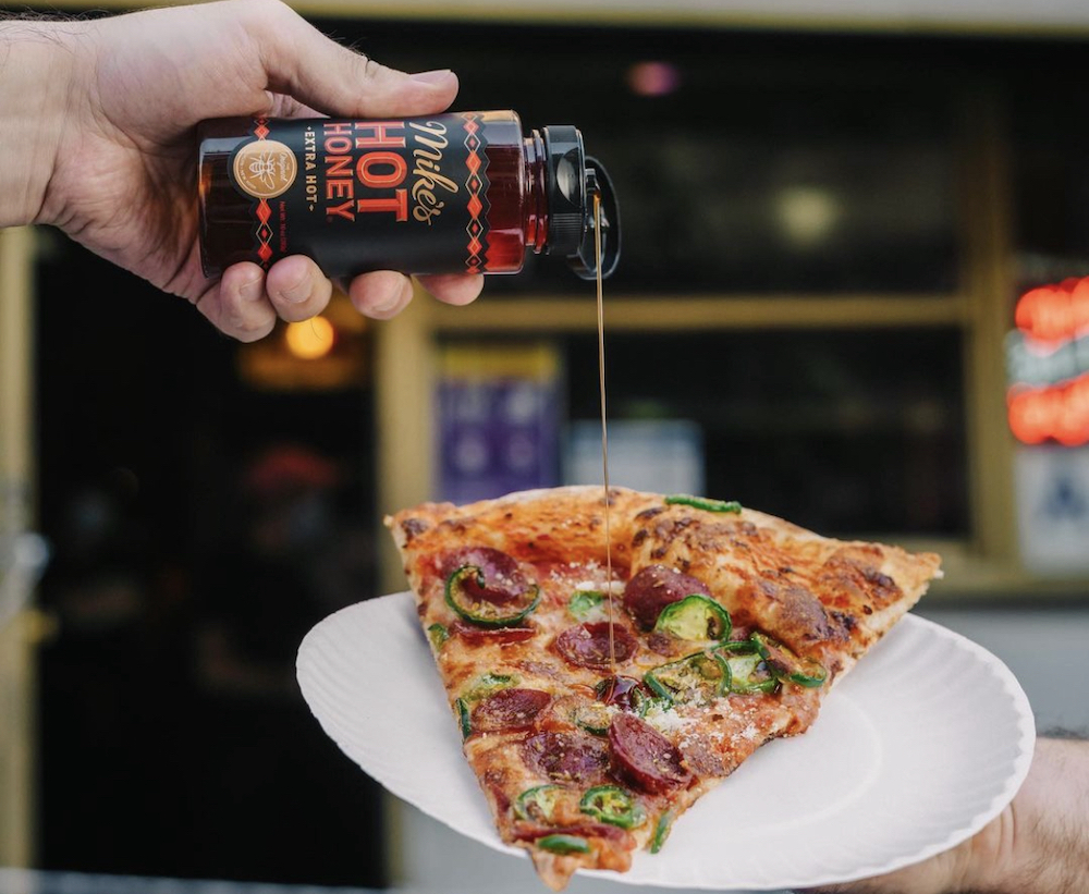 This photo shows a man's hand holding a bottle of Mike's Hot Honey and dripping it over a pizza slice topped with beef pepperoni and jalapeños