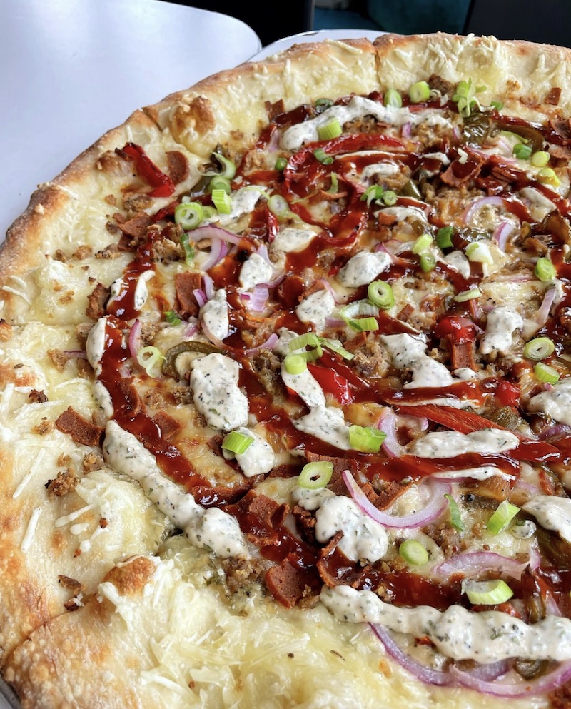 This photo shows the BBQ Supreme pizza from Screamers Pizzeria, topped with vegan bacon, vegan sausage, red onions, peppers, scallions and BBQ sauce