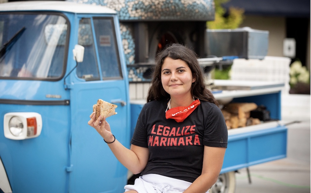 This photo shows Sofia Arango, with long dark hair and a T-shirt that says Legalize Marinara, eating a slice of pizza in front of Atlanta Pizza Truck's trademark blue food truck.