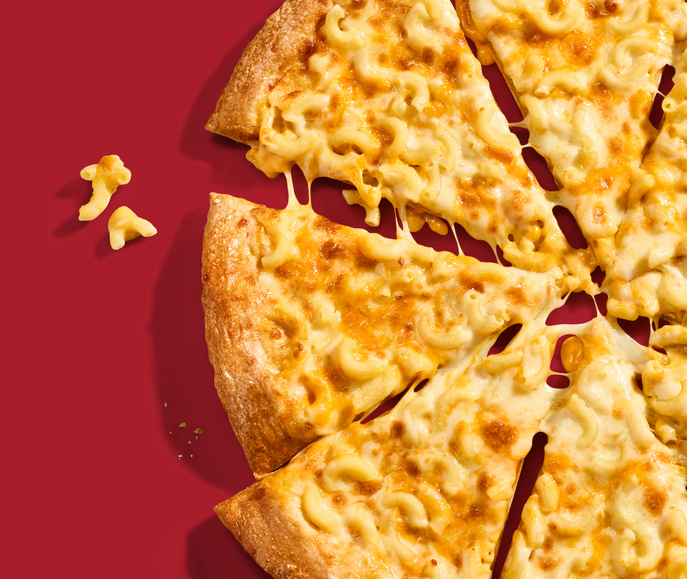 This photo shows half of a whole pizza topped with macaroni and cheese and three stray pieces of macaroni to the side, all set against a dark red background.