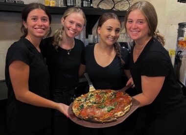 This photo shows four female servers, all wearing black t-shirts, holding one large pizza and smiling for the camera.