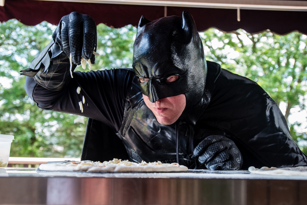 This photo shows Jim in a Batman costume, leaning in close and adding cheese to an unbaked pizza.